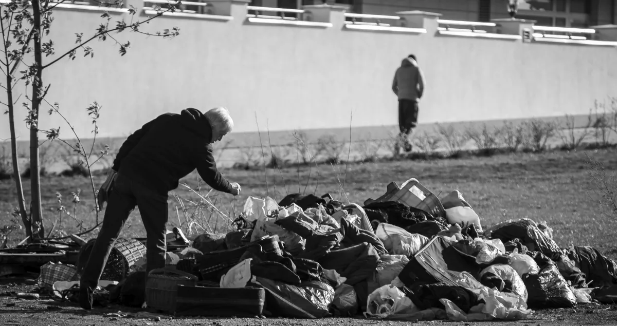 Man searching through a heap of discarded clothes while another figure walks away behind him